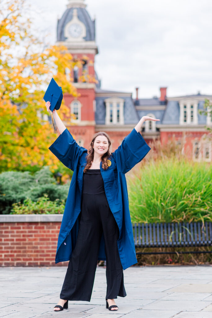wvudecembergradphotos12 carrie white photography graduate in a blue cap and gown cheers by tossing her cap in the air on a campus with red brick buildings and autumn trees in the background