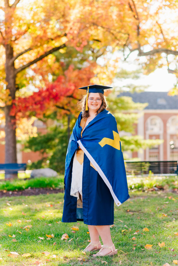 wvudecembergradphotos13 carrie white photography graduate in blue cap and gown with yellow tassel stands on a grassy campus area amid fall foliage holding a blue and gold flag draped over shoulders