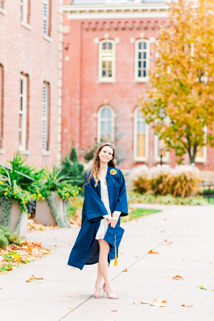 wvudecembergradphotos18 carrie white photography graduate in a blue gown and cap stands on a campus path with autumn leaves and brick buildings in the background posing for a photo
