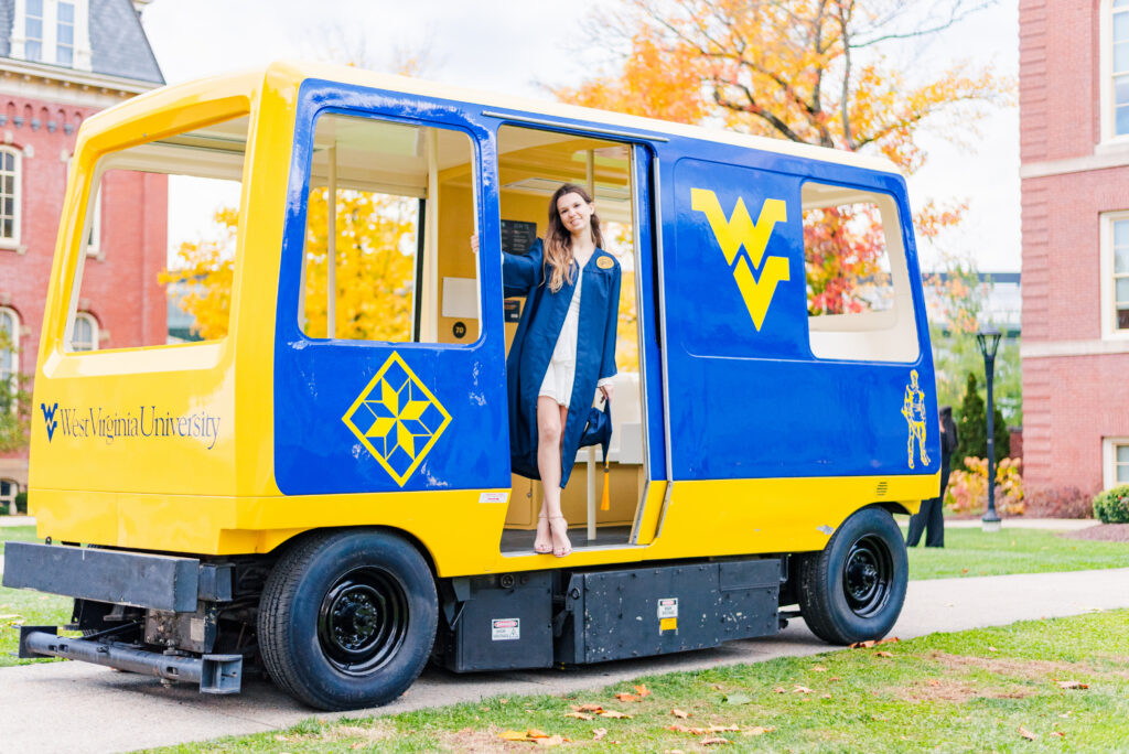 student in a blue coat steps off a yellow and blue west virginia university campus shuttle with the wv logo on the side and autumn trees in the background