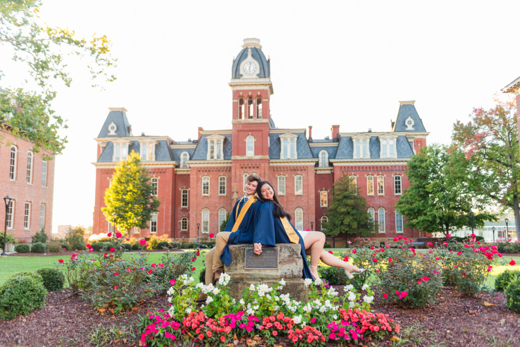 wvudecembergradphotos23 carrie white photography couple in blue graduation gowns sitting on a pedestal in a rose garden with a red brick campus building and clock tower behind them