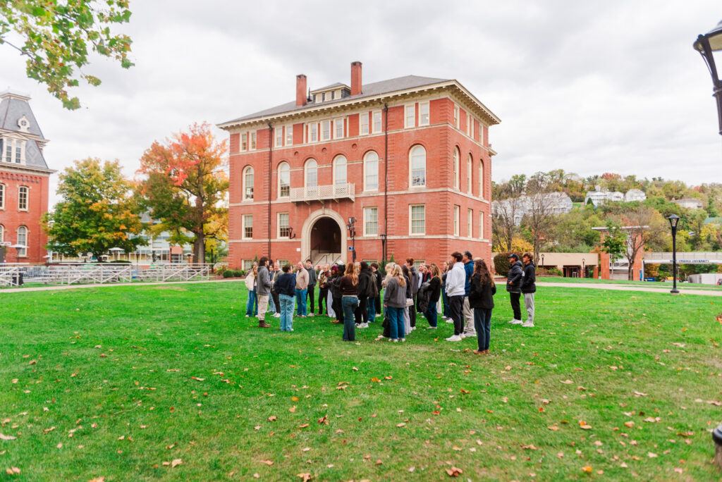 wvudecembergradphotos24 carrie white photography group of students gathered on a green lawn in front of a red brick college building with arched entrance