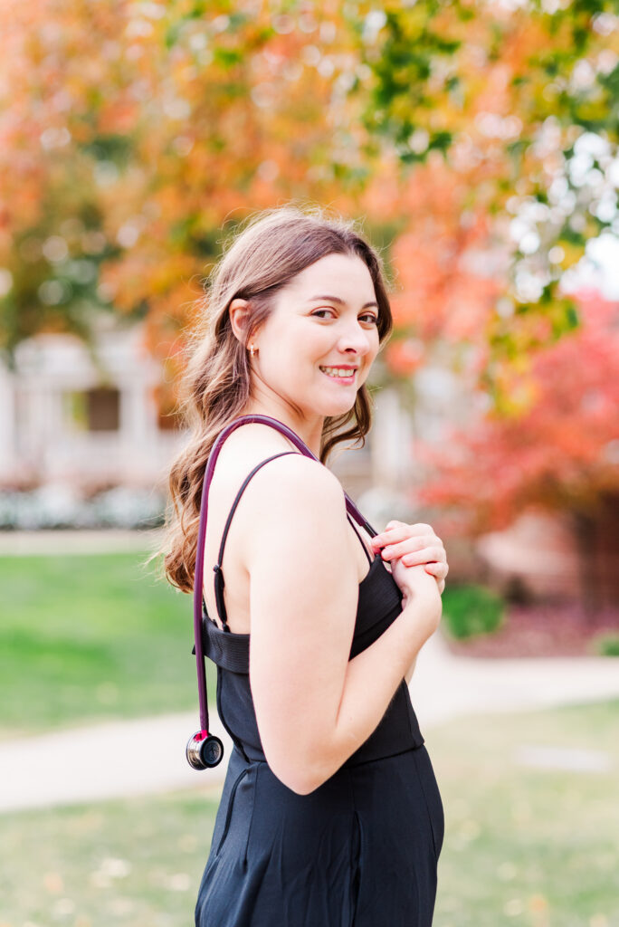wvudecembergradphotos25 carrie white photography smiling woman in a black dress posing outdoors with colorful autumn leaves in the background