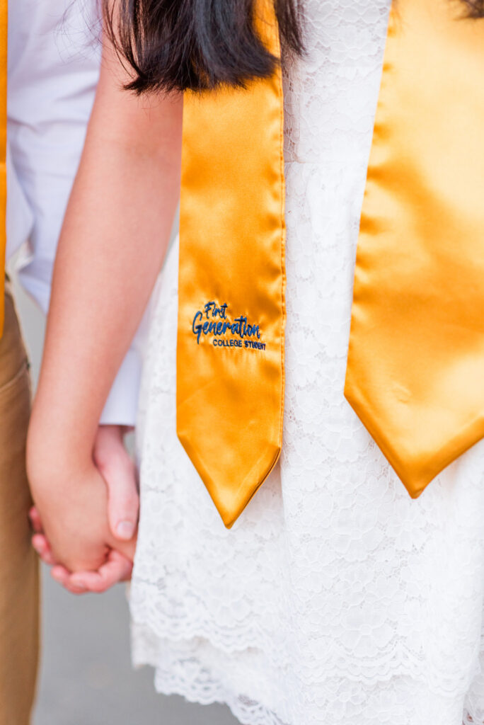 wvudecembergradphotos29 carrie white photography close up of two graduates holding hands wearing orange sashes over white lace dresses one sash reads'First Generation College Student'.