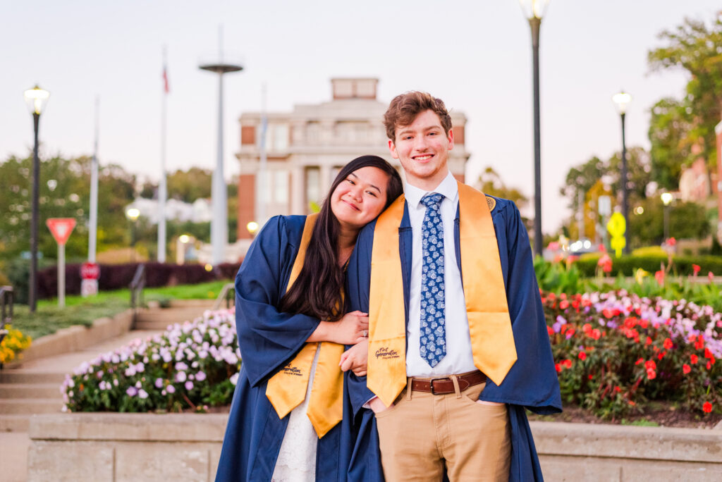 wvudecembergradphotos34 carrie white photography two graduates in blue gowns and gold stoles smile together outdoors on campus with flowers and a building in the background
