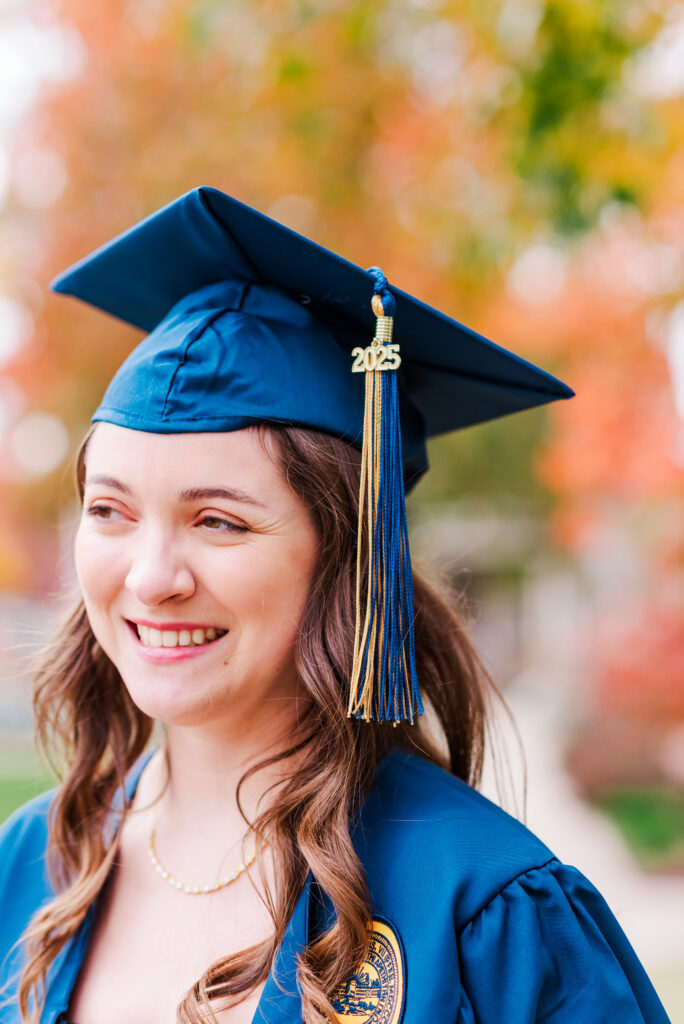 wvudecembergradphotos37 carrie white photography smiling graduate in blue cap and gown with gold tassel marked autumn background during wvu december graduation photos