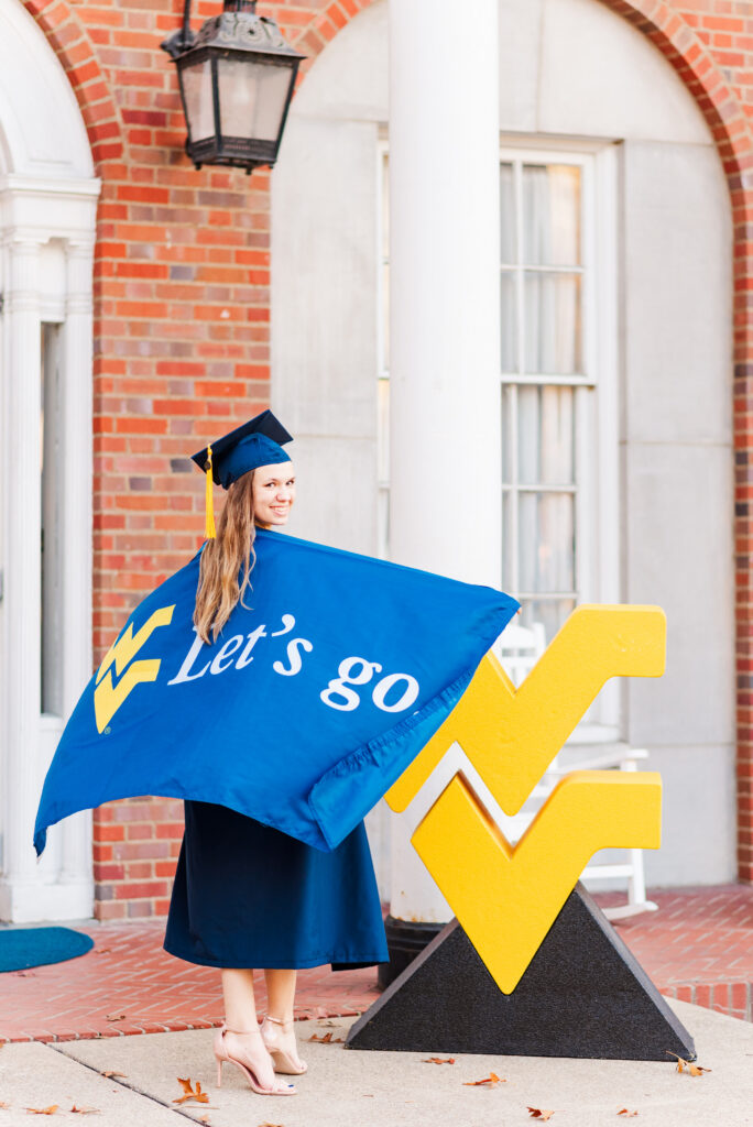 wvudecembergradphotos39 carrie white photography graduate in blue cap and gown posing with a blue banner that reads'Let's go', outdoors beside a large yellow chevron sculpture.