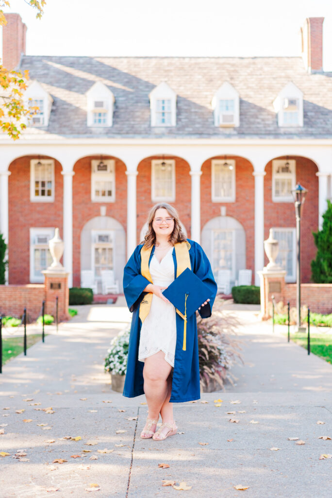 wvudecembergradphotos48 carrie white photography graduate in a blue cap and gown holding a diploma on a campus walkway with a red brick building in the background