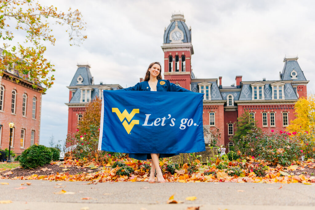 wvudecembergradphotos50 carrie white photography smiling woman in a blue graduation gown holding a blue banner that reads'Let's go' on a university campus with red brick buildings and autumn leaves in the foreground.