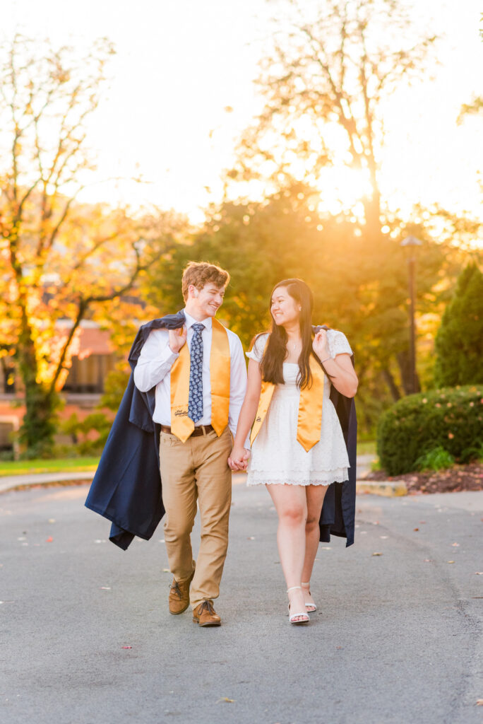 wvudecembergradphotos58 carrie white photography couple in graduation gowns walking hand in hand outdoors at sunset smiling at each other