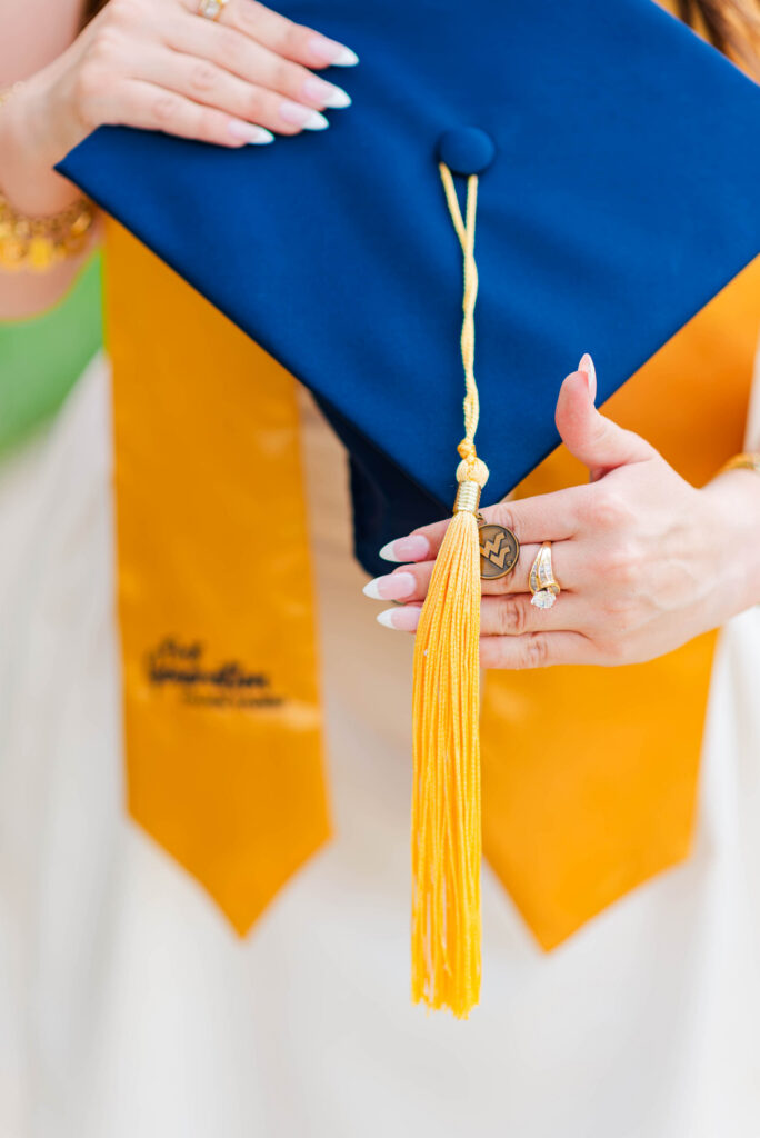 wvuevansdalecampusgraduationphotos02 carrie white photography close up of a navy graduation cap with a yellow tassel held by a graduate wearing an orange gown and rings on the fingers