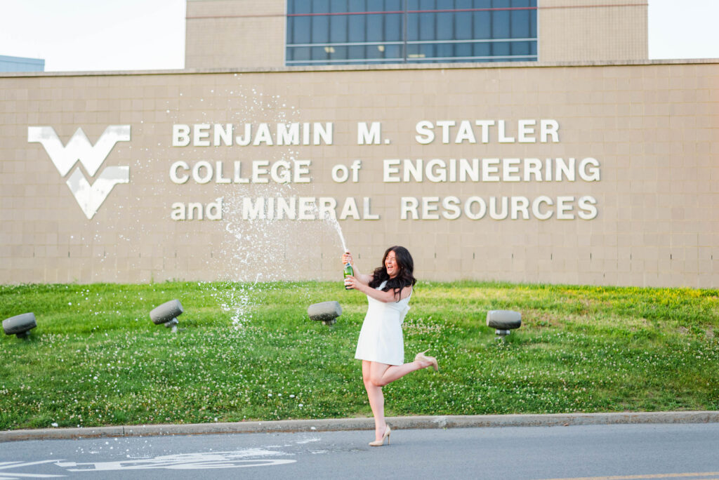 woman in a white dress joyfully spraying champagne outdoors in front of a brick building with large engineering college signage in the background during wvu evansdale graduation photos