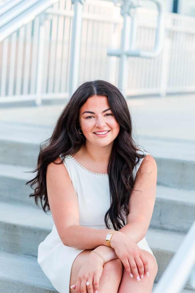smiling woman with long dark hair in a white dress sitting on outdoor stairs wearing pearl necklace and gold jewelry