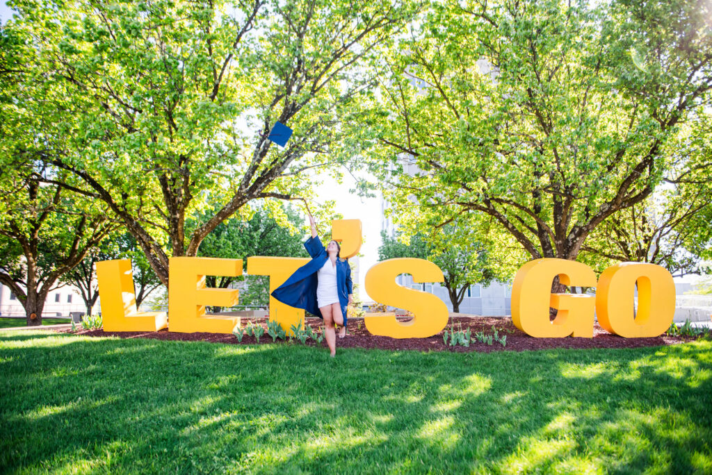 graduate in a blue graduation gown stands among oversized yellow letters outdoors in a sunny campus park