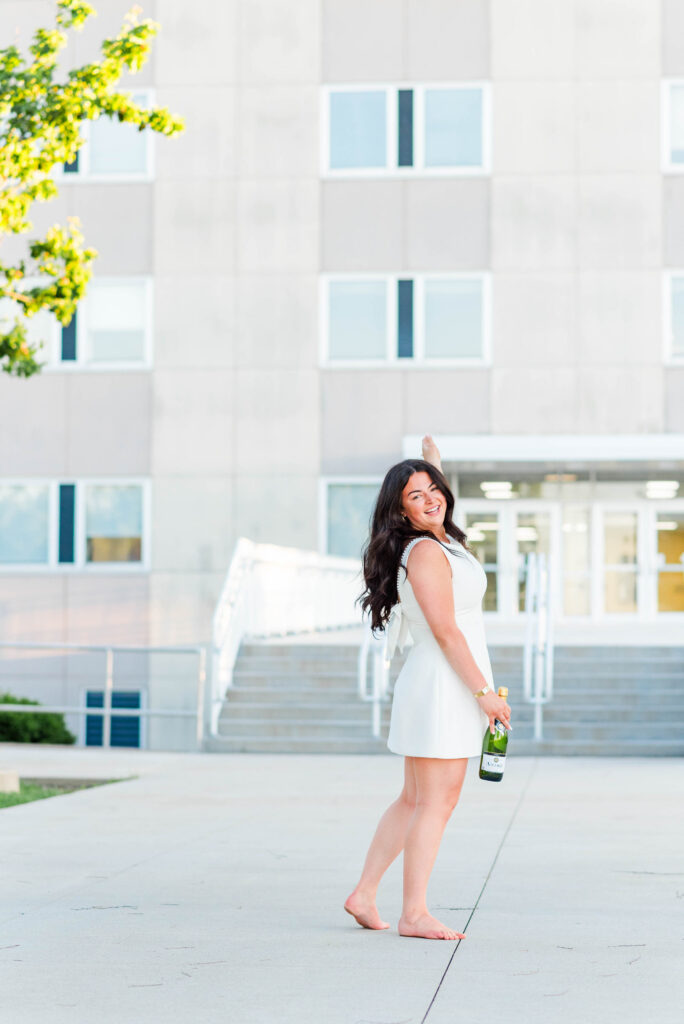 smiling woman in a white dress barefoot holding a champagne bottle outdoors with a modern building in the background