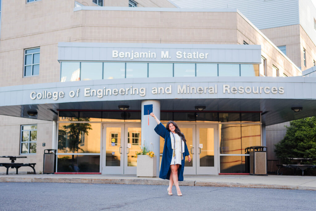 graduate in a blue gown posing in front of the benjamin m statler college of engineering and mineral resources entrance