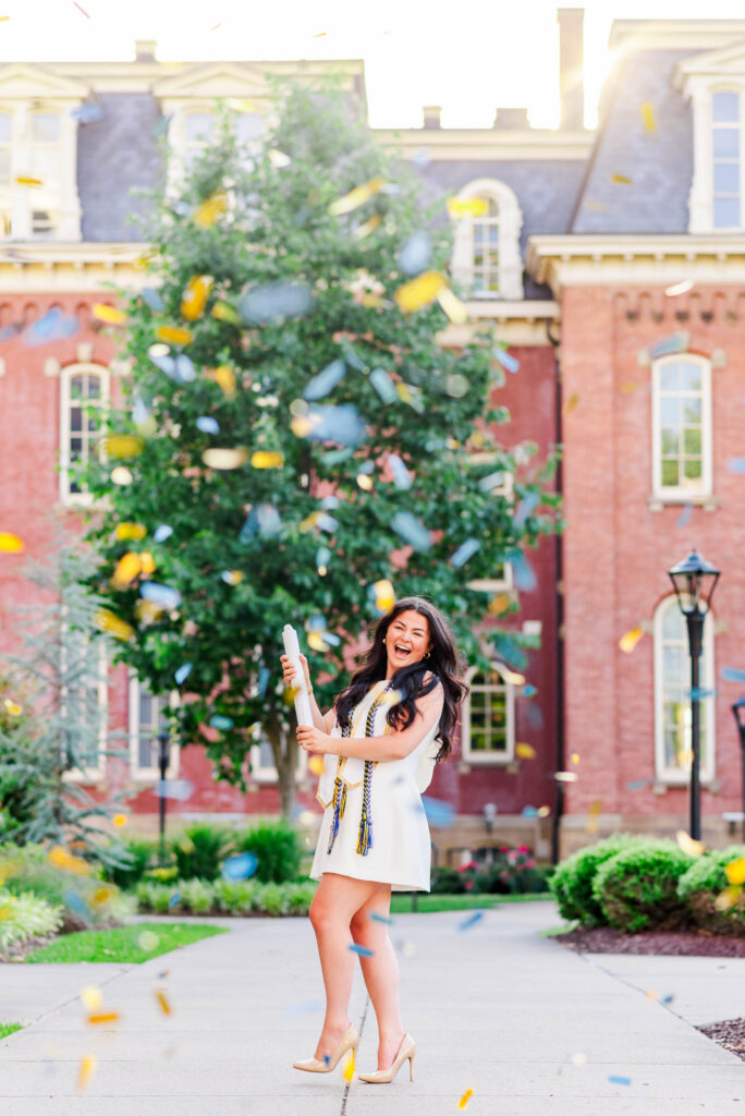 wvuevansdalecampusgraduationphotos15 carrie white photography woman in a white dress holds a diploma and smiles while confetti falls in an outdoor courtyard