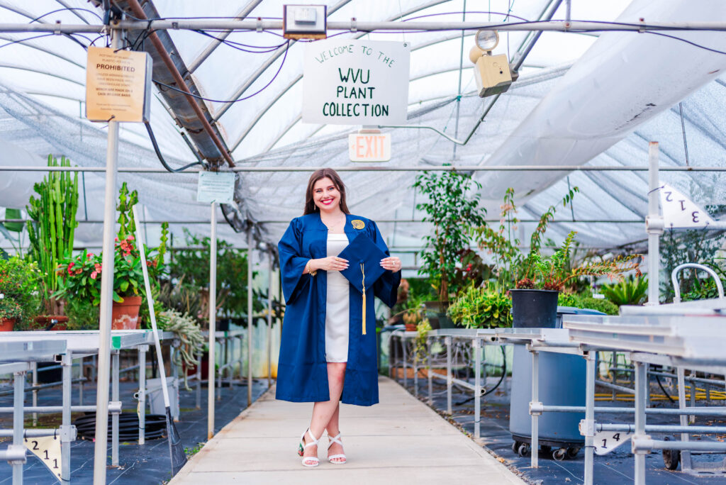 graduate in a blue gown stands in a greenhouse smiling holding a diploma under a'WVU Plant Collection' sign during WVU Evansdale graduation photos
