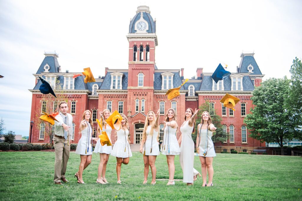 wvugradphotos carrie white photography graduates in white dresses tossing yellow and blue caps on a campus lawn in front of a red brick clock tower building