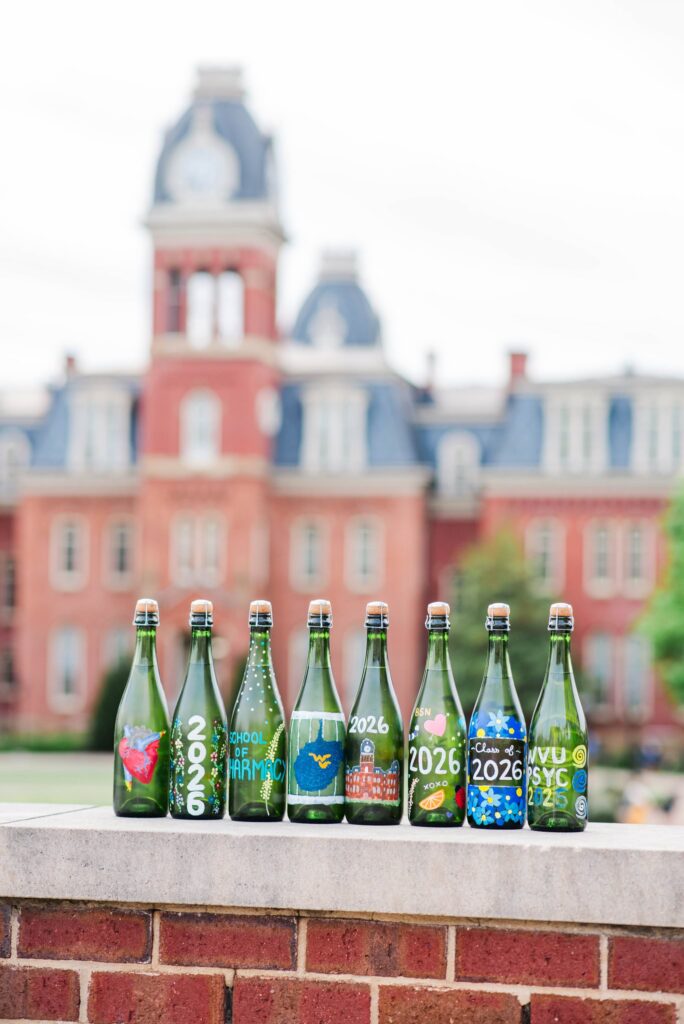 row of green glass bottles with colorful school themed labels lined up on a stone ledge a blurred campus building in the background