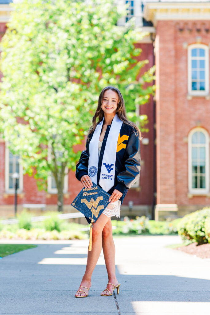 wvu grad posing with decorated graduation cap and wearing gymnastics letterman jacket during wvu grad photos