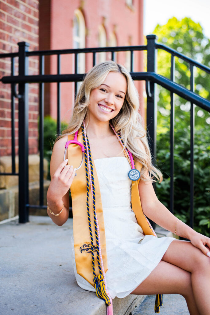 smiling young graduate in a white dress with a yellow stole and cords sitting on outdoor steps beside a brick building