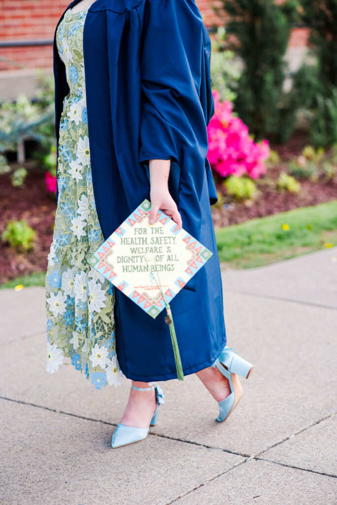 graduate in a blue gown holding a decorative sign about health safety welfare and dignity wearing a floral dress and teal heels in a garden