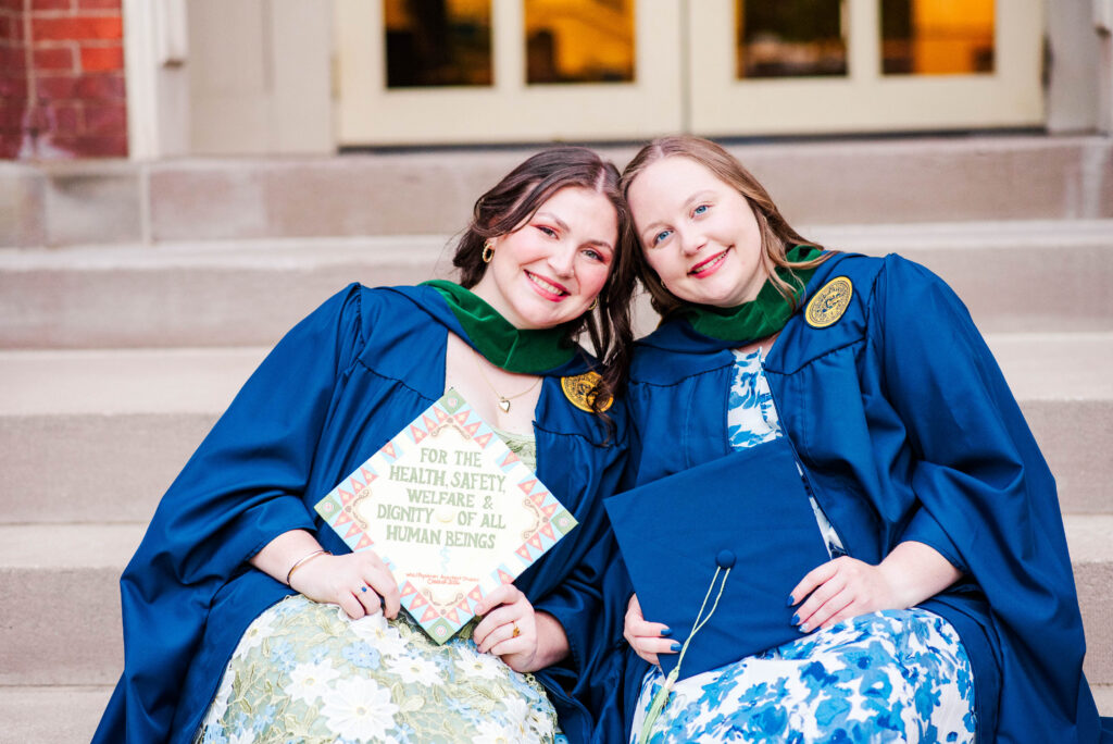 two graduates in blue gowns sit on stone steps smiling at the camera with one holding a colorful poster or program