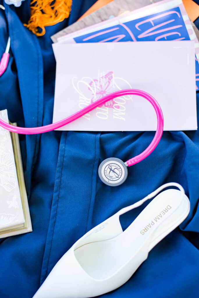 pink stethoscope resting on blue scrubs beside notebooks and a white medical shoe