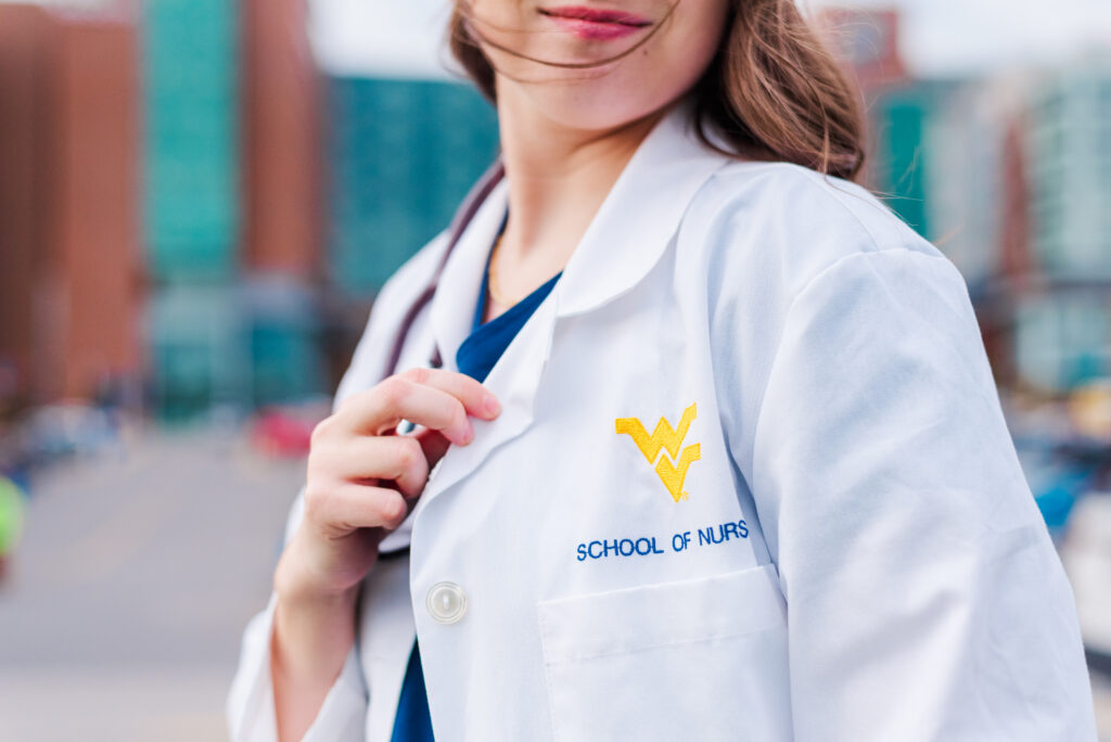 nurse student in a white coat with a yellow wv logo adjusting the lapel outdoors text'SCHOOL OF NURS' visible).