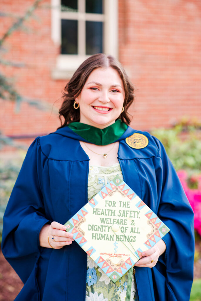 smiling graduate in a blue gown with a green hood outdoors holding a colorful diploma cover that reads'For the health, safety, welfare & dignity of all human beings'.