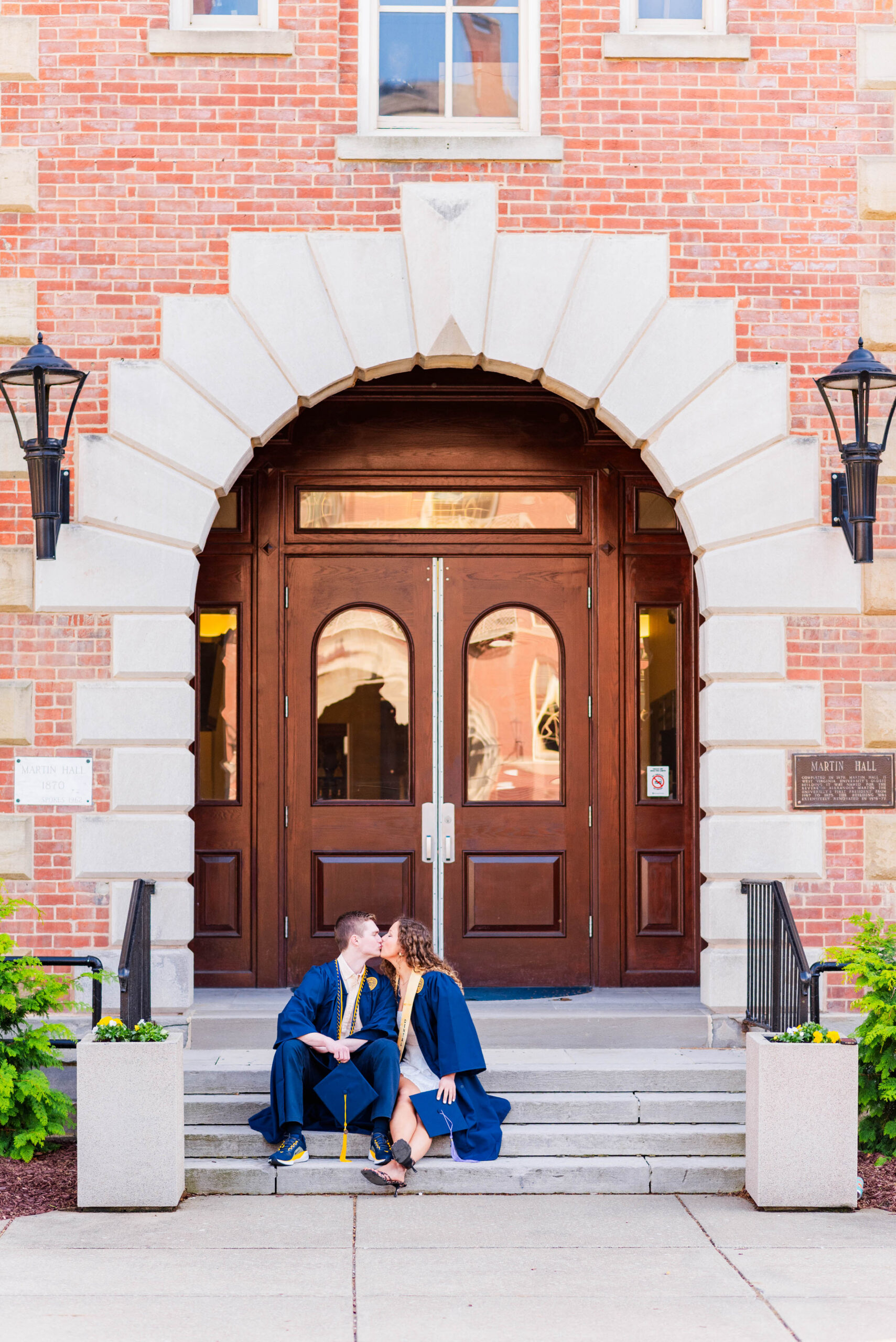 two graduates in blue gowns sit on steps and kiss in front of a red brick campus entrance with a large arched doorway