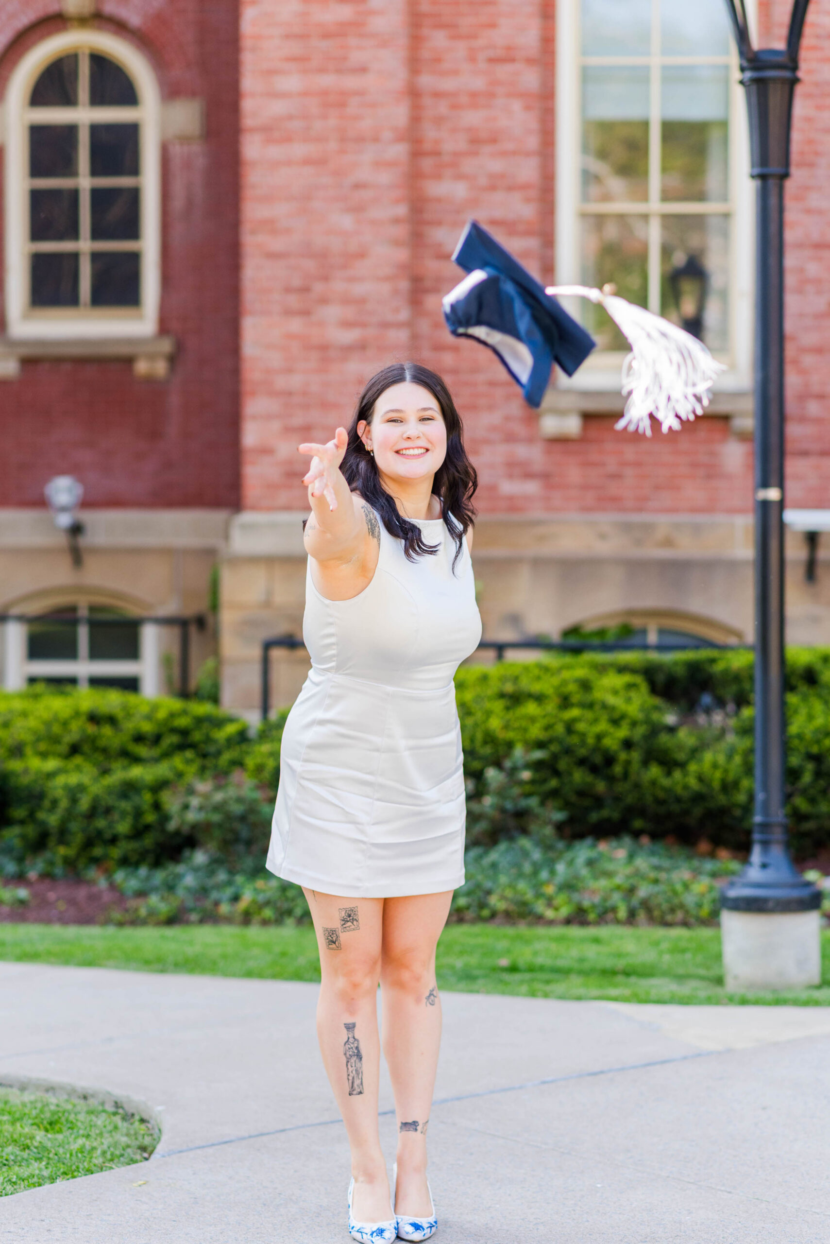 woman in a white dress throws a blue graduation cap in the air outside a brick campus building smiling joyfully