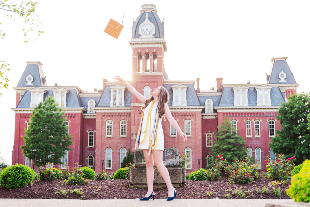 graduationcaptoss1 carrie white photography wvu grad in white dress tossing cap up in the air in front of woodburn hall in morgantown wv during sunset