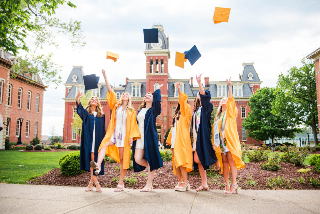 graduationcaptoss5 carrie white photography a group of wvu grads wearing blue and yellow graduation gowns all tossing graduation caps in the air at the same time in front of woodburn hall in morgantown wv